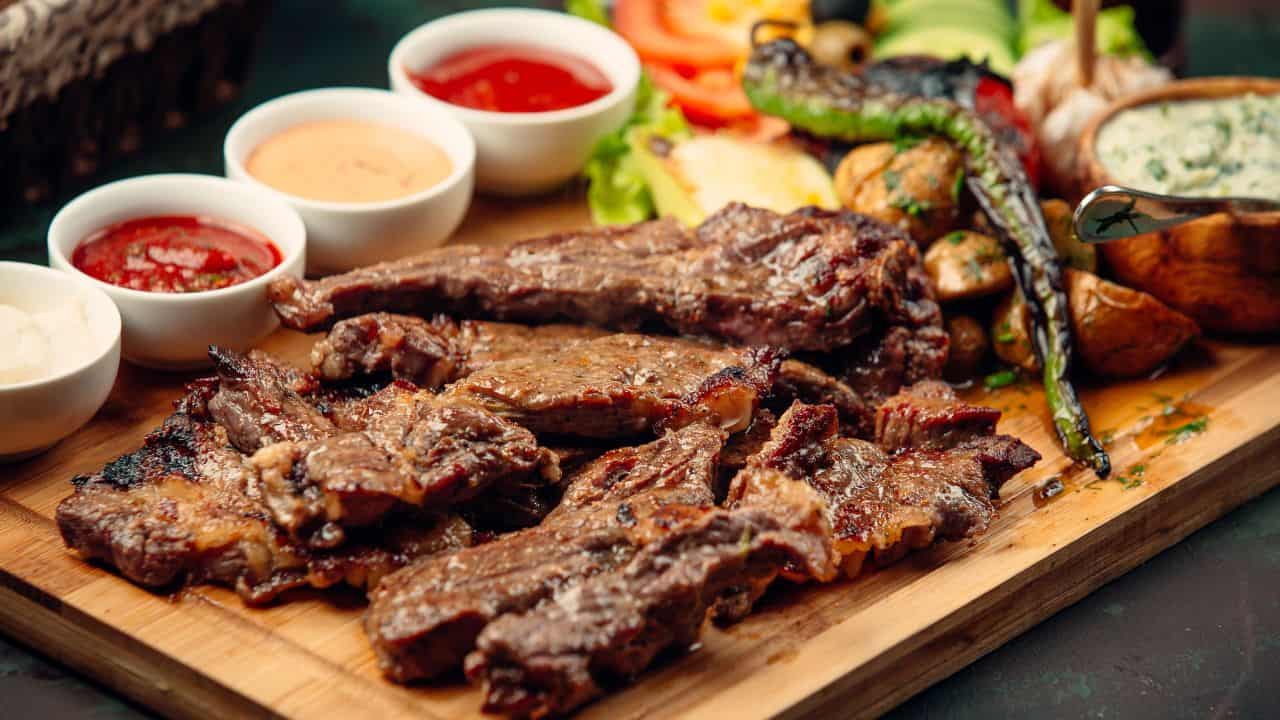 Grilled steak slices on a wooden board, served with assorted grilled vegetables and four small bowls of dipping sauces.