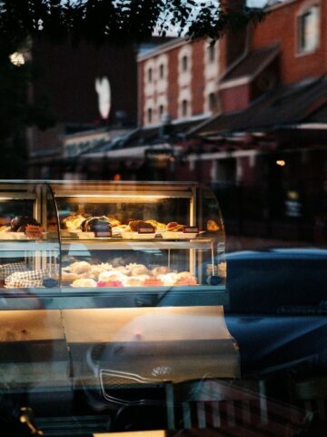 A lit pastry display case with various desserts is seen through a window at dusk, with reflections of the street and buildings visible in the glass.