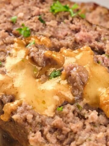 A slice of meatloaf with melted cheese in the center, garnished with chopped parsley, served alongside mashed potatoes.