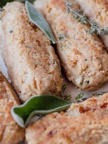 Six baked, golden-brown vegan sausages are arranged on a plate, garnished with fresh sage leaves and thyme sprigs.