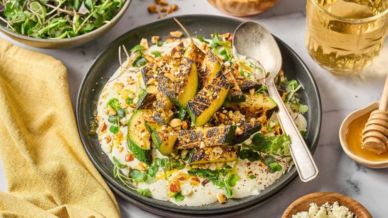 Grilled zucchini slices served on a bed of yogurt with microgreens, chopped nuts, and honey, accompanied by a spoon, a glass of drink, and a side salad.