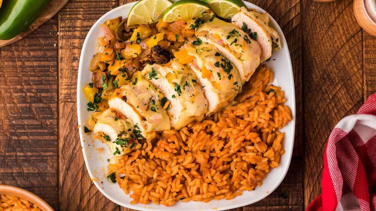 A plate with sliced chicken breast, sautéed vegetables, lime wedges, and a serving of seasoned rice on a wooden table.
