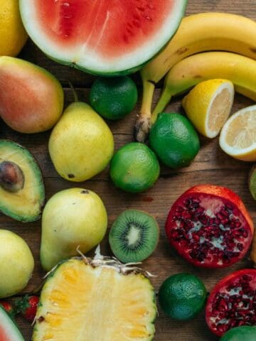 An assortment of fresh fruits including watermelon, bananas, avocados, pears, lemons, limes, strawberries, grapes, pineapple, pomegranate, and kiwi arranged on a wooden surface.