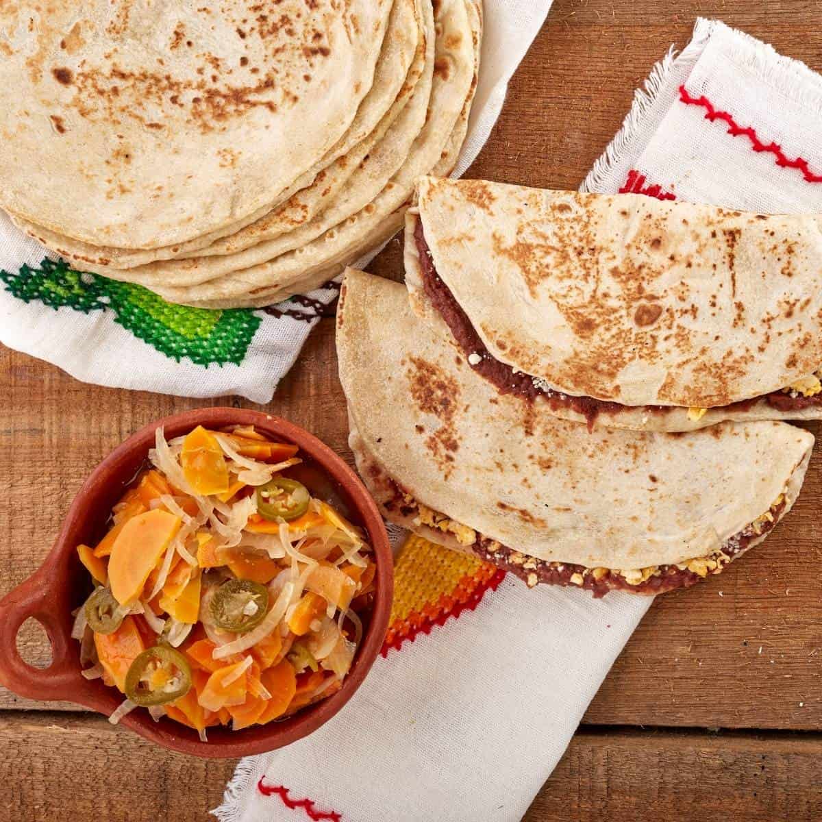 A stack of tortillas and two folded tortillas filled with beans and cheese—a classic Honduran food—sit on a cloth next to a bowl of pickled vegetables on a wooden surface.