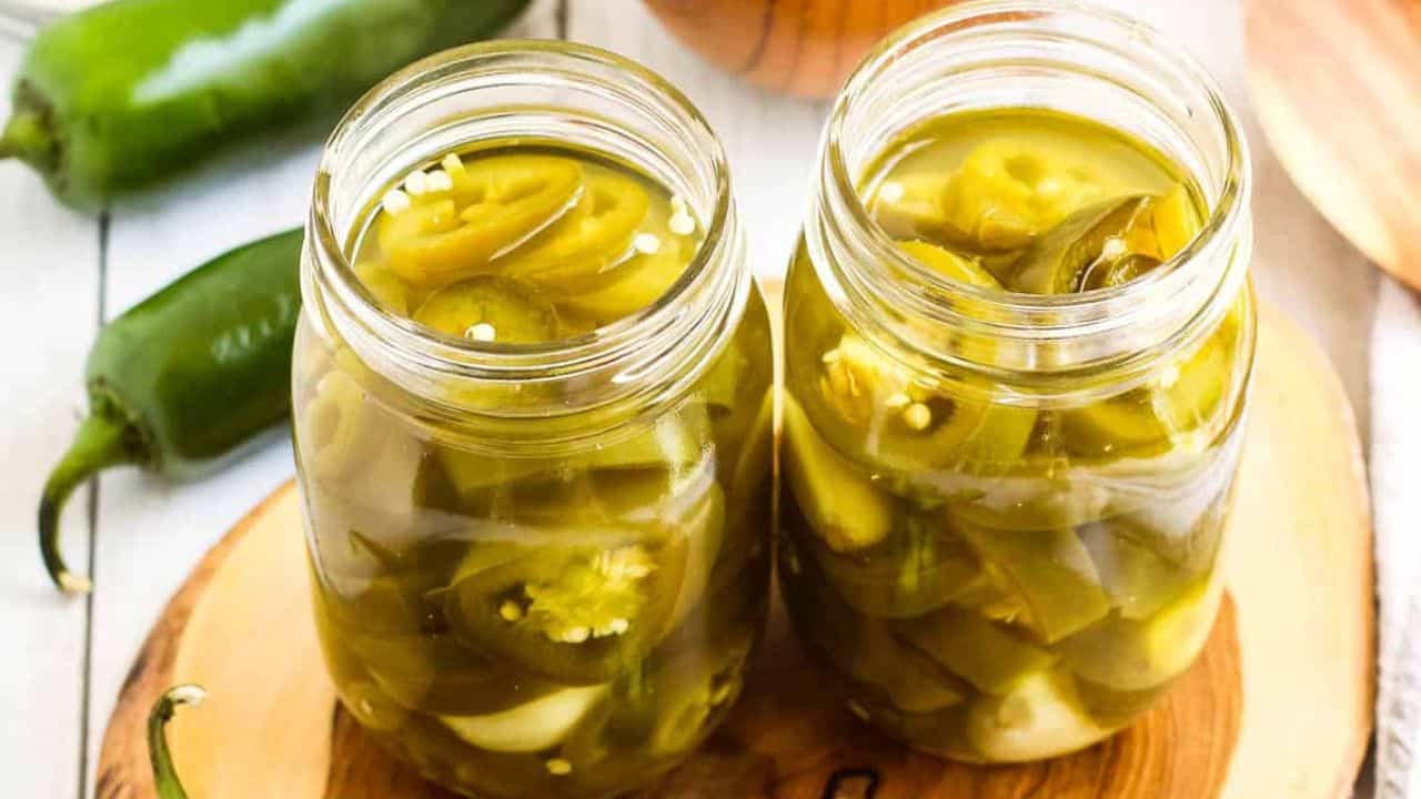 Two canning jars filled with pickled jalapenos on a wooden board with fresh jalapenos on the side.