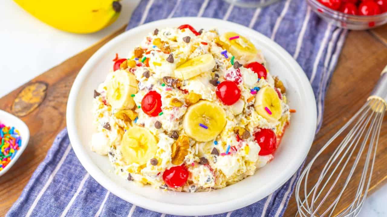 A bowl of banana split salad topped with banana slices, maraschino cherries, walnuts, chocolate chips, and colorful sprinkles sits on a plate with a whisk and bananas nearby.