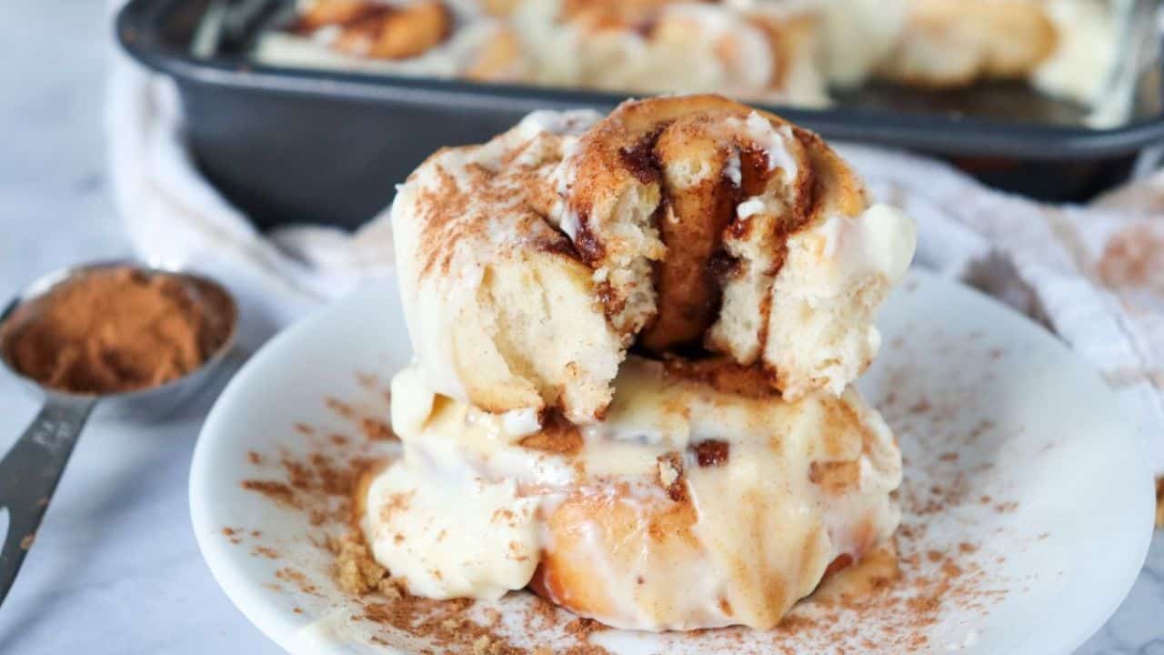 A close-up of a cinnamon roll with white icing being lifted from a pan filled with more iced cinnamon rolls.