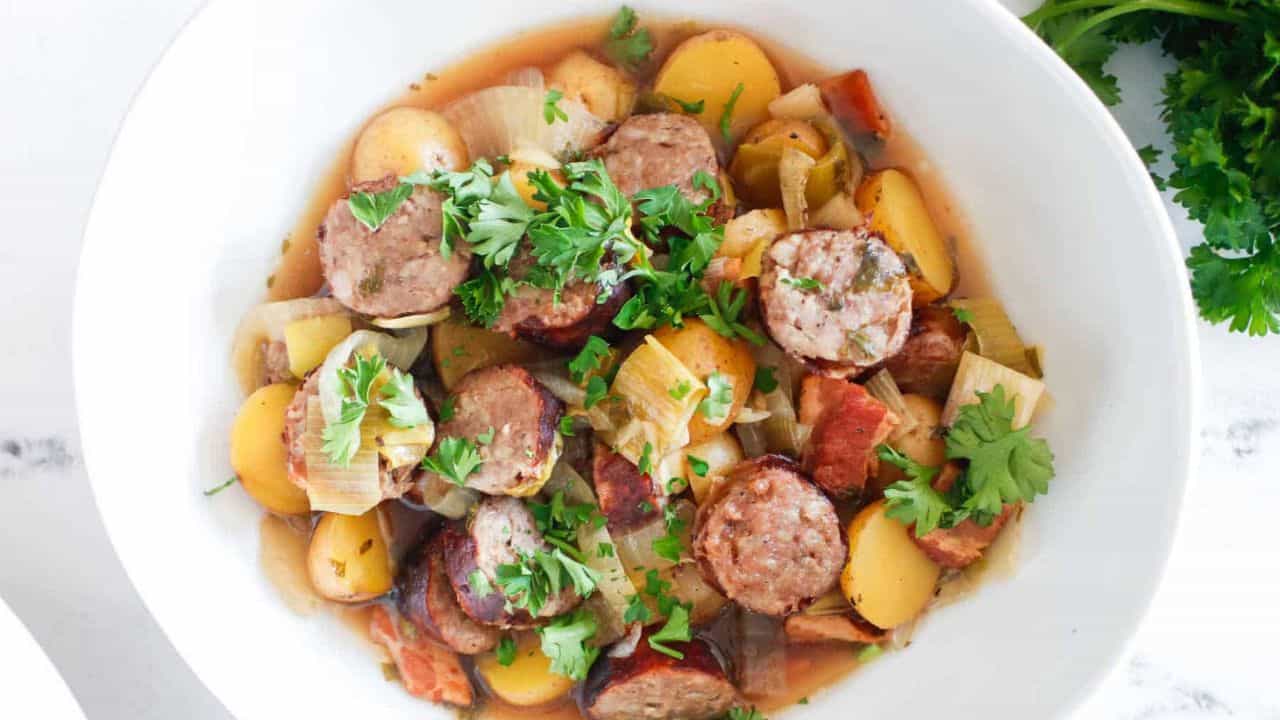 Sliced sausages with cooked onions, potatoes, and vegetables in a white baking dish.