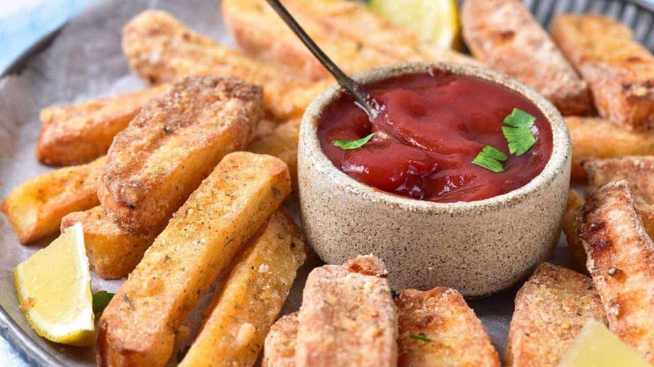 A plate of thick, crispy fries arranged around a small bowl of red dipping sauce, garnished with parsley, with lemon wedges on the side.