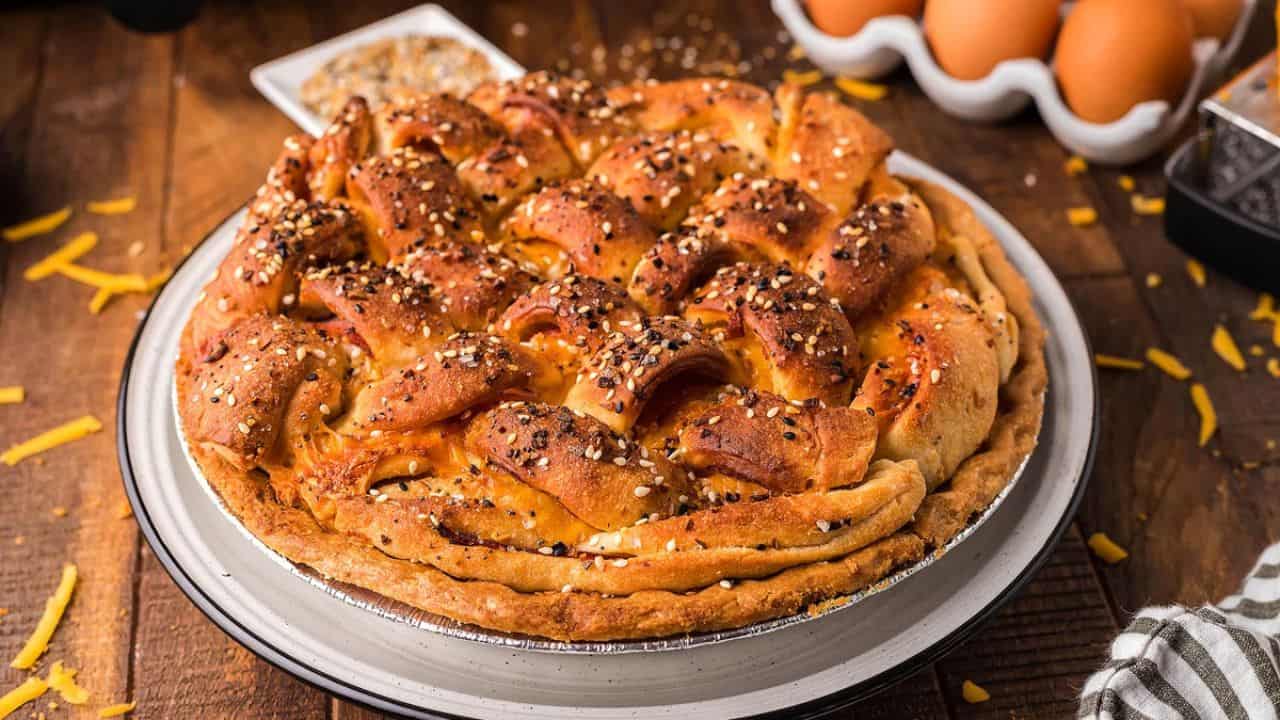 A baked pie with a golden, lattice-style crust topped with seeds sits on a white plate; eggs and shredded cheese are in the background.