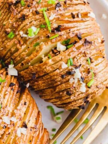 Two hasselback potatoes garnished with chopped chives, garlic, and coarse salt, served on a white plate with a gold fork.