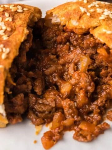 A baked bun with a golden crust and sesame seeds is split open, revealing a savory filling of ground beef and diced vegetables on a white plate.