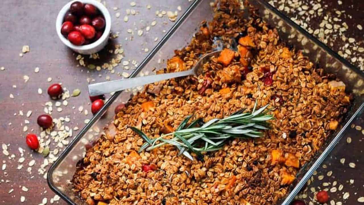 A baking dish filled with a baked oat and fruit mixture, topped with a sprig of rosemary. A spoon rests in the dish. Oats and cranberries are scattered on the table, and a small bowl of cranberries is visible in the background.