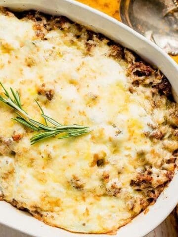 A baked casserole topped with melted cheese and garnished with a sprig of rosemary, next to a silver spoon on a textured yellow cloth.