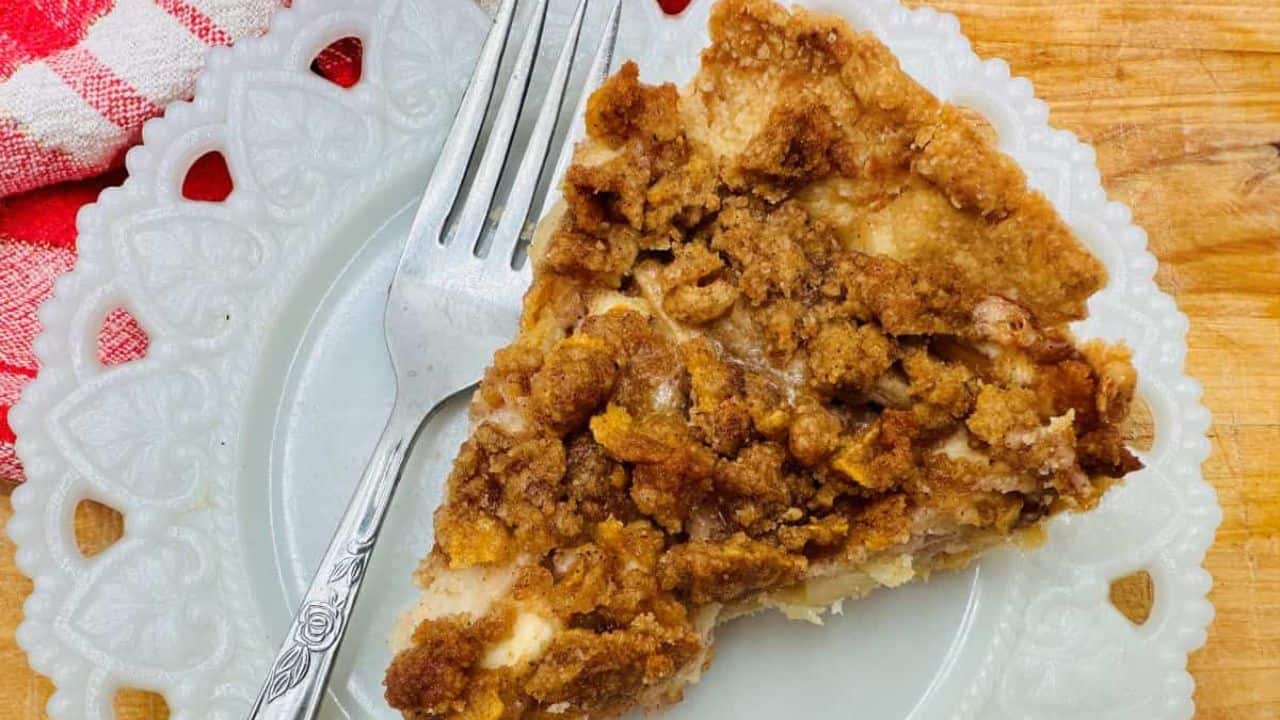 A slice of crumb-topped pie on a white decorative plate with a fork next to it. A red and white cloth is partially visible in the corner.