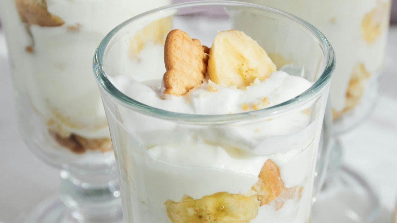 Glass of layered dessert with whipped cream, banana slices, and cookie pieces, with similar desserts visible in the background.