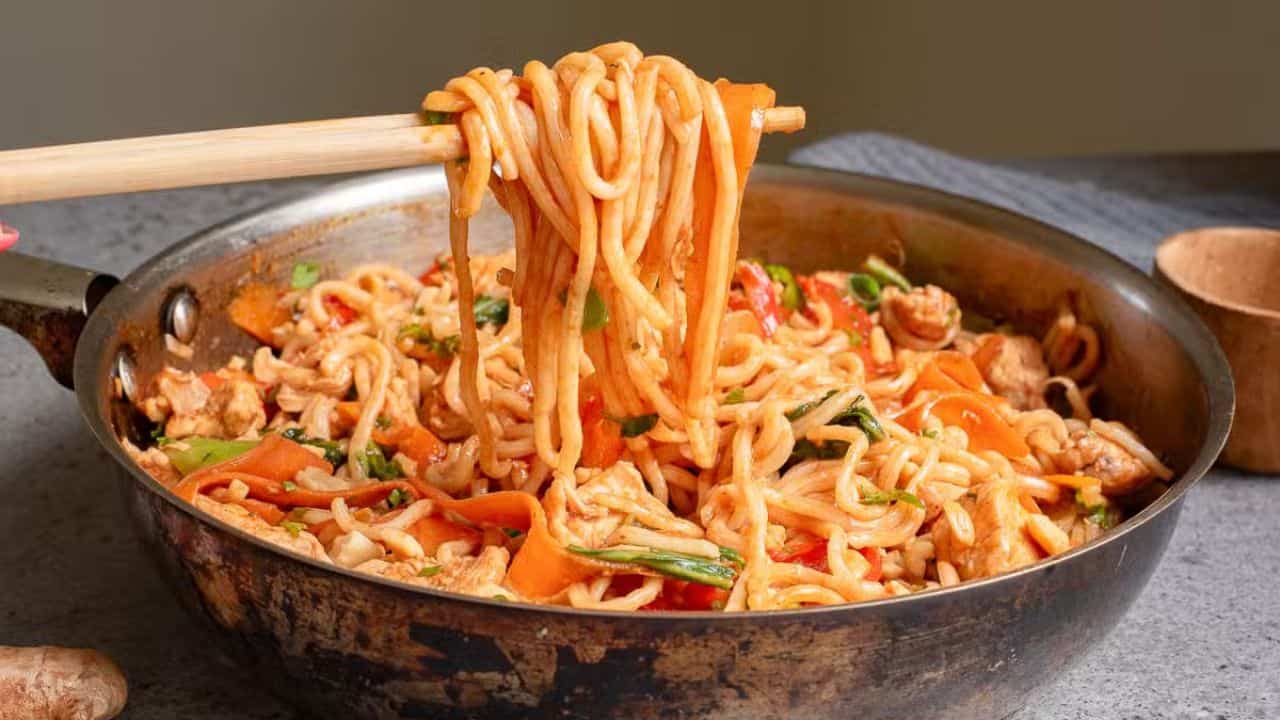 A bowl of noodle stir-fry topped with vegetables, chicken, crushed peanuts, and fresh mint leaves. Chopsticks rest on the side of the bowl, which is placed on a textured surface with a small container in the background.