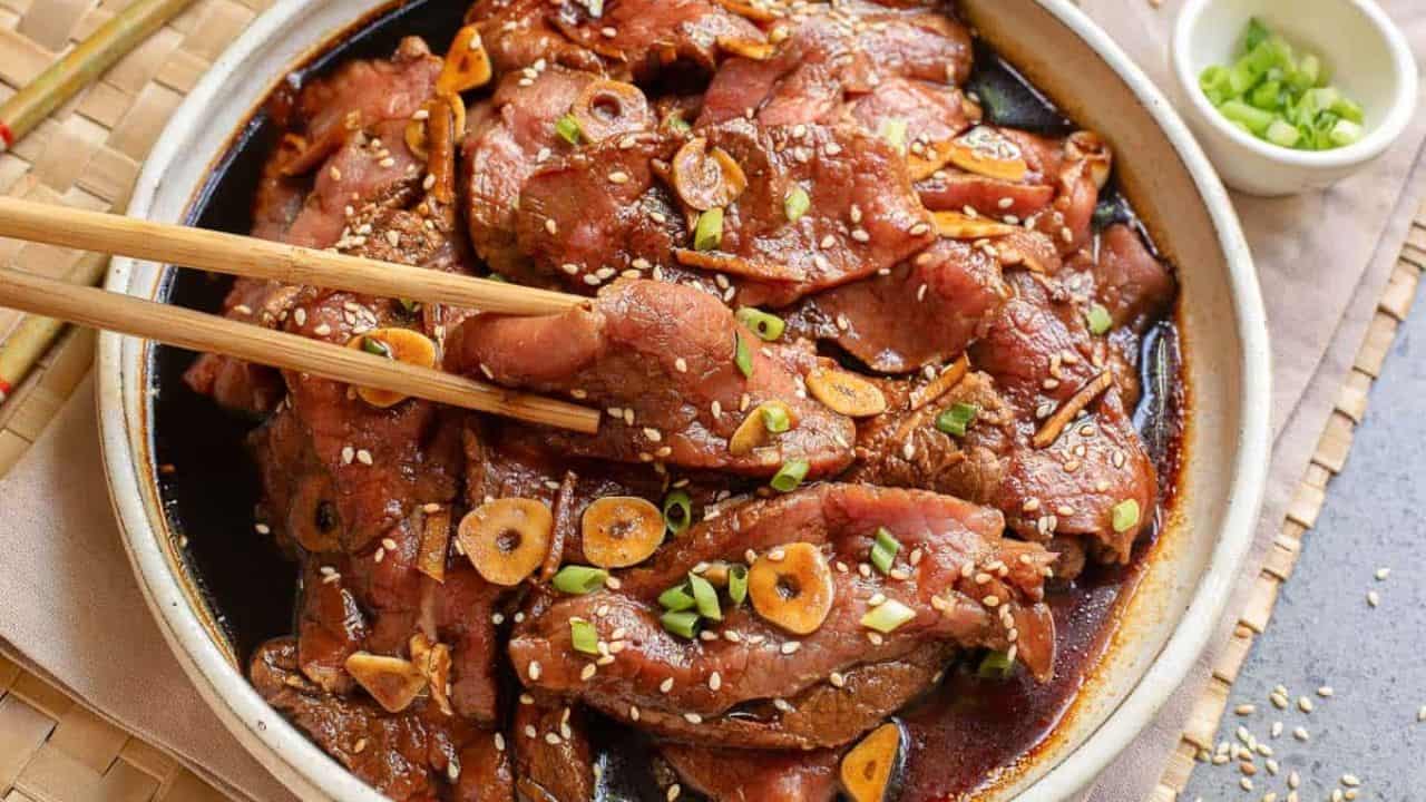 A plate of marinated grilled beef topped with sliced garlic, sesame seeds, and chopped green onions, with chopsticks picking up a piece.