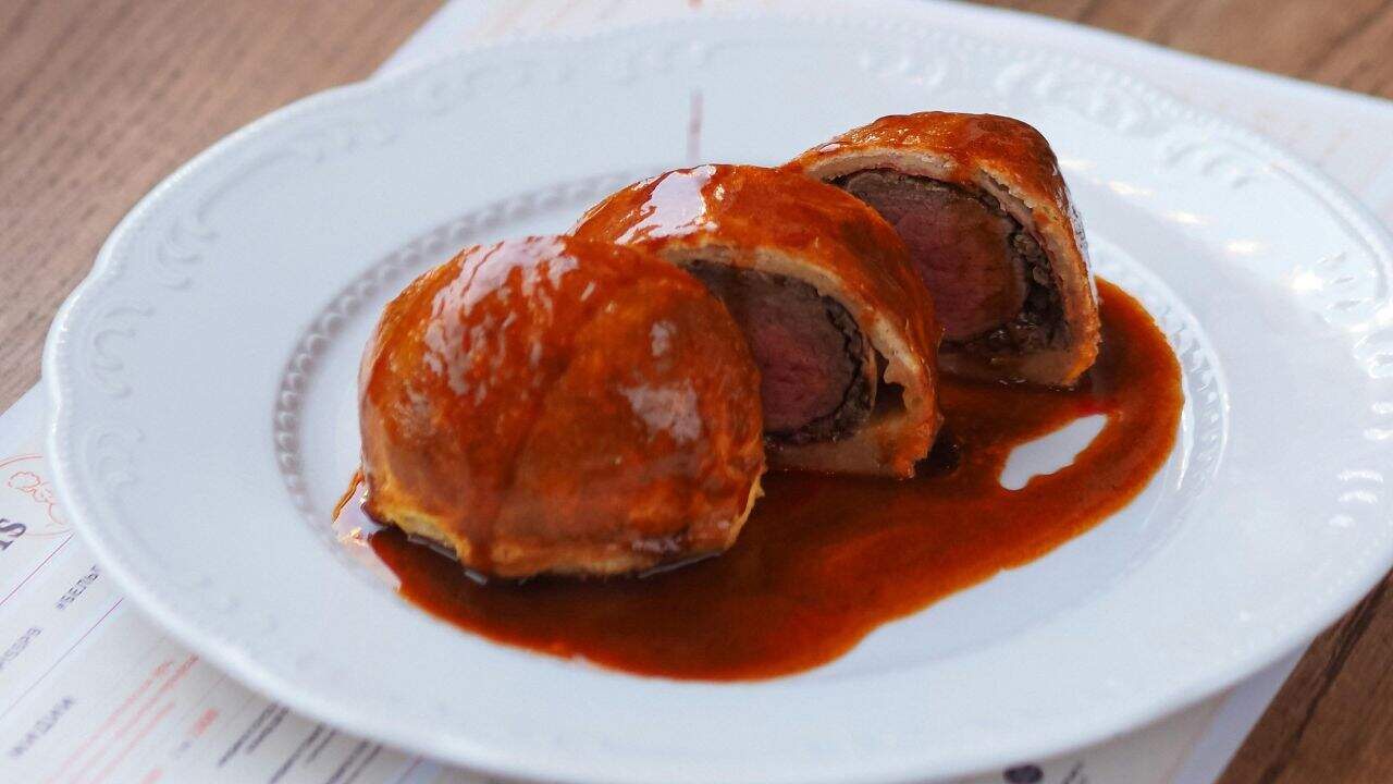 Two slices of beef Wellington with golden pastry are served on a white plate, topped with a glossy brown sauce.