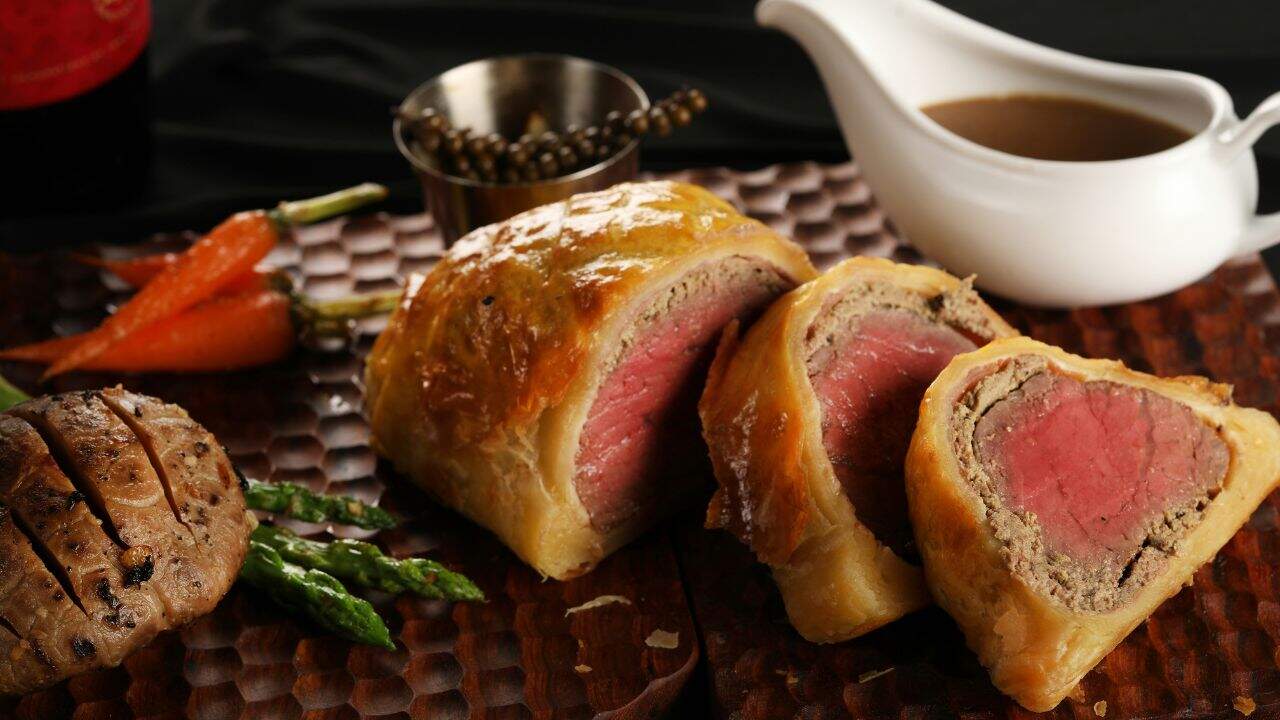 Sliced beef Wellington with medium-rare center, served on a wooden board with grilled vegetables, peppercorns, and a sauce boat.
