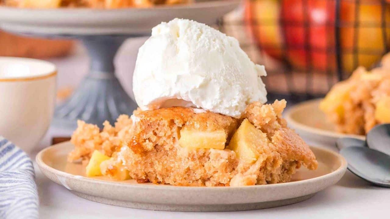 A slice of apple cake topped with a scoop of vanilla ice cream on a plate, with more dessert and apples in the background.