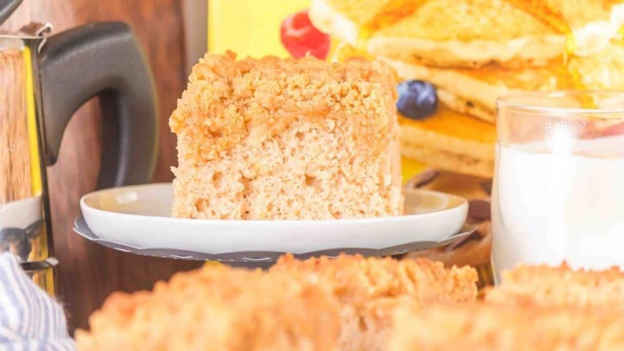 A slice of crumb cake on a white plate, with a glass of milk and a stack of pancakes in the background.