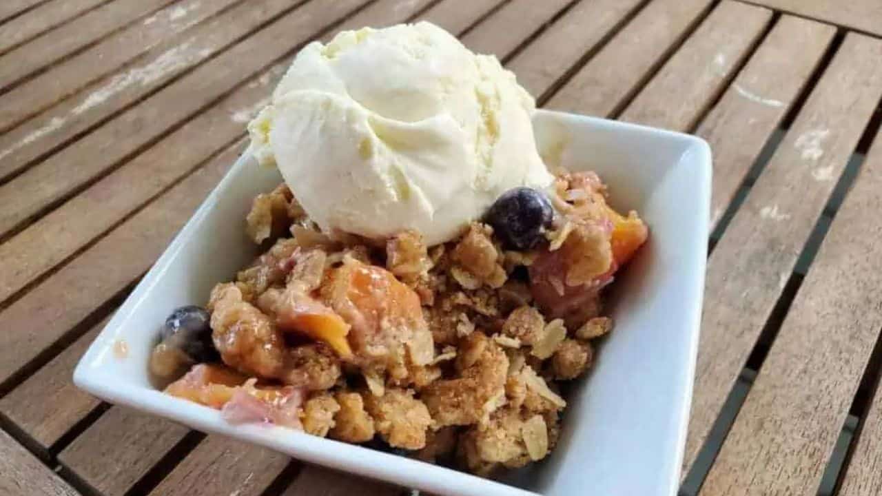 Image shows a white bowl with a serving of blueberry peach crisp topped with a scoop of ice cream and some whipped cream.