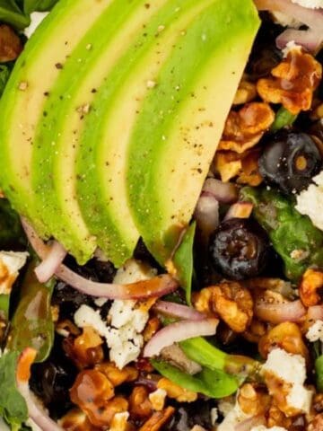 Close-up of a salad with avocado slices, walnuts, blueberries, red onion, spinach, and crumbled cheese in a white bowl.