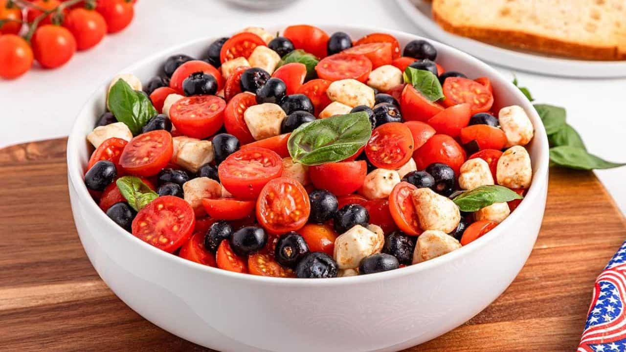 A white bowl filled with cherry tomatoes, black olives, mozzarella balls, and fresh basil leaves sits on a wooden board.