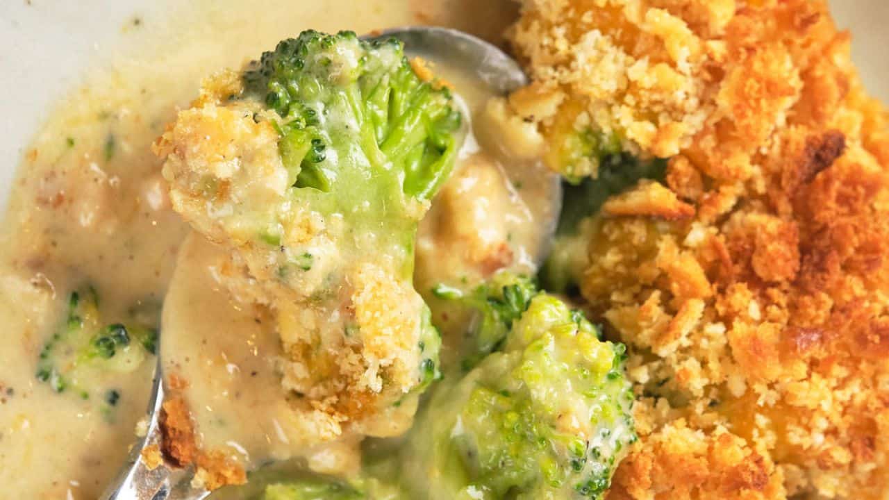 A close-up of broccoli casserole with a creamy sauce and a golden-brown breadcrumb topping, served in a white dish with a spoon.