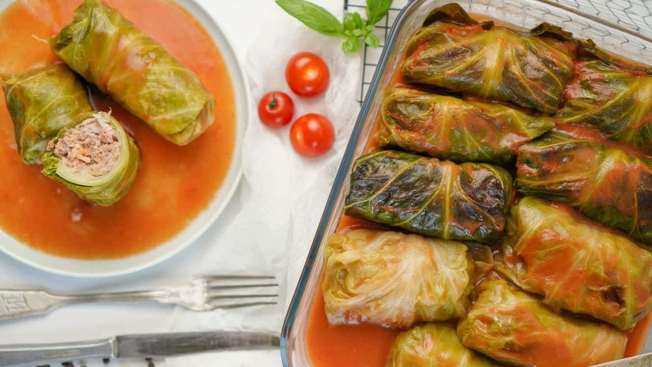 Cabbage rolls filled with meat and rice in tomato sauce, served on a plate and in a baking dish, with cherry tomatoes and basil on the side.