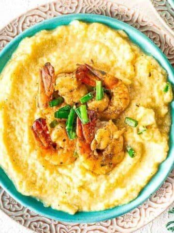 A bowl of shrimp and grits topped with chopped green onions, served on a patterned plate next to a green napkin and seasoning bowls.