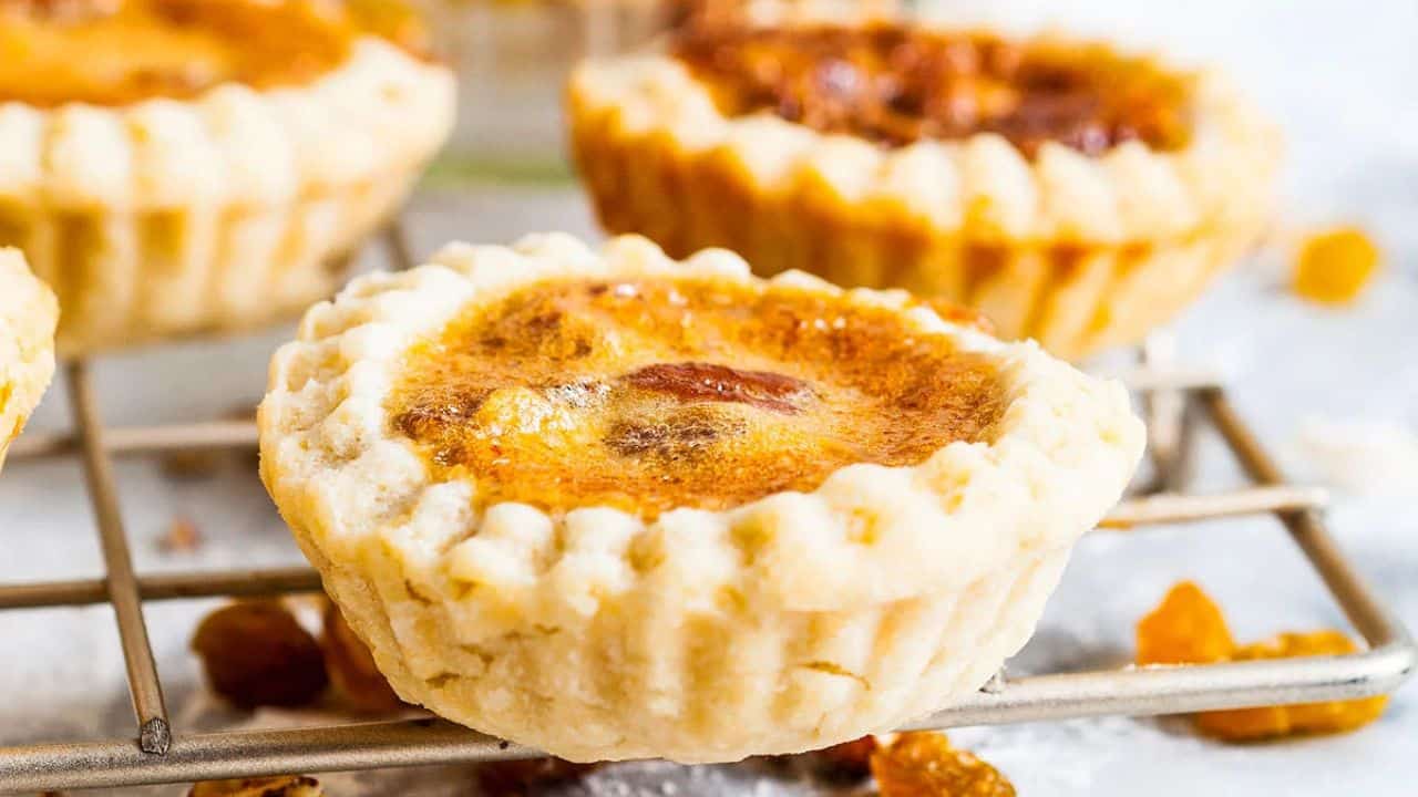 Close-up of a butter tart with a golden, flaky crust and a caramelized filling, sitting on a metal cooling rack with a few raisins nearby.