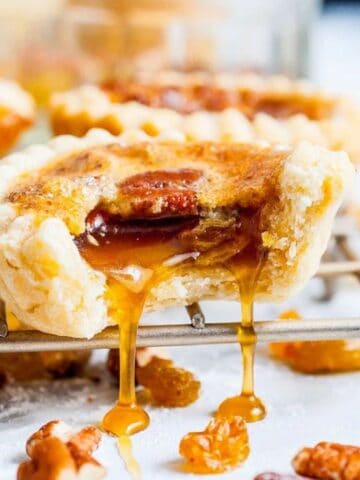 A close-up of a butter tart with a bite taken out, showing gooey filling and pecans, resting on a cooling rack with scattered nuts and raisins.