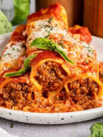 Three baked cannelloni pasta tubes filled with ground meat, topped with tomato sauce, grated cheese, and fresh basil, served on a white plate with a fork.