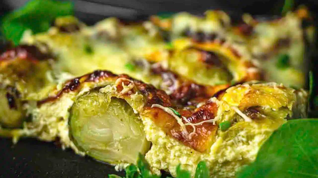 Close-up of a baked dish featuring Brussels sprouts in a creamy, cheesy sauce with a golden-brown crust, served with fresh greens.
