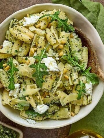 A bowl of pasta with pesto sauce, arugula, ricotta cheese, and pine nuts, surrounded by smaller plates of pasta, pesto, pine nuts, and pieces of bread on a table.