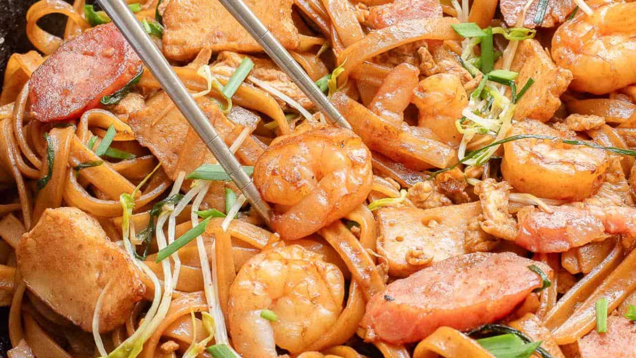 A close-up of stir-fried flat noodles with shrimp, sliced sausage, and vegetables, served on a banana leaf.