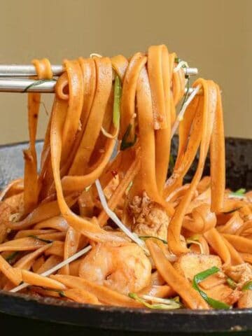 A hand holds chopsticks lifting stir-fried flat noodles with shrimp, vegetables, and meat from a black skillet.