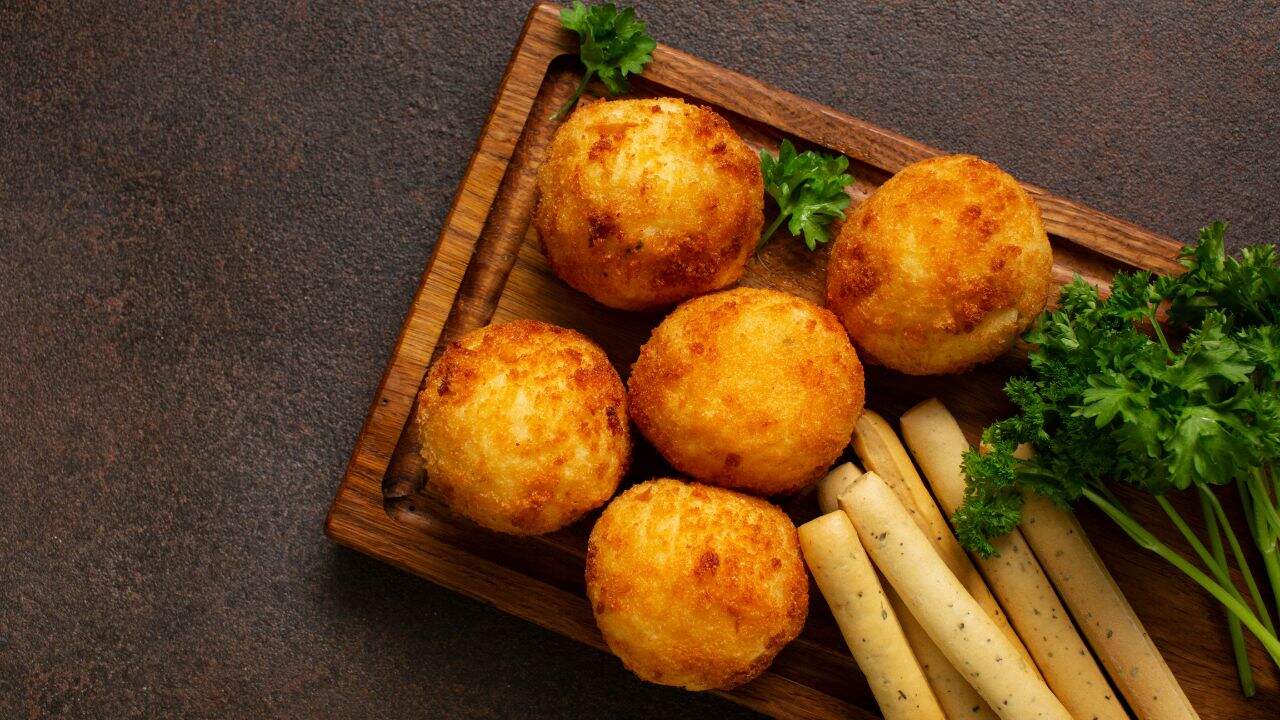 Five golden-brown fried cheese balls and several breadsticks are arranged on a wooden tray, garnished with fresh parsley.