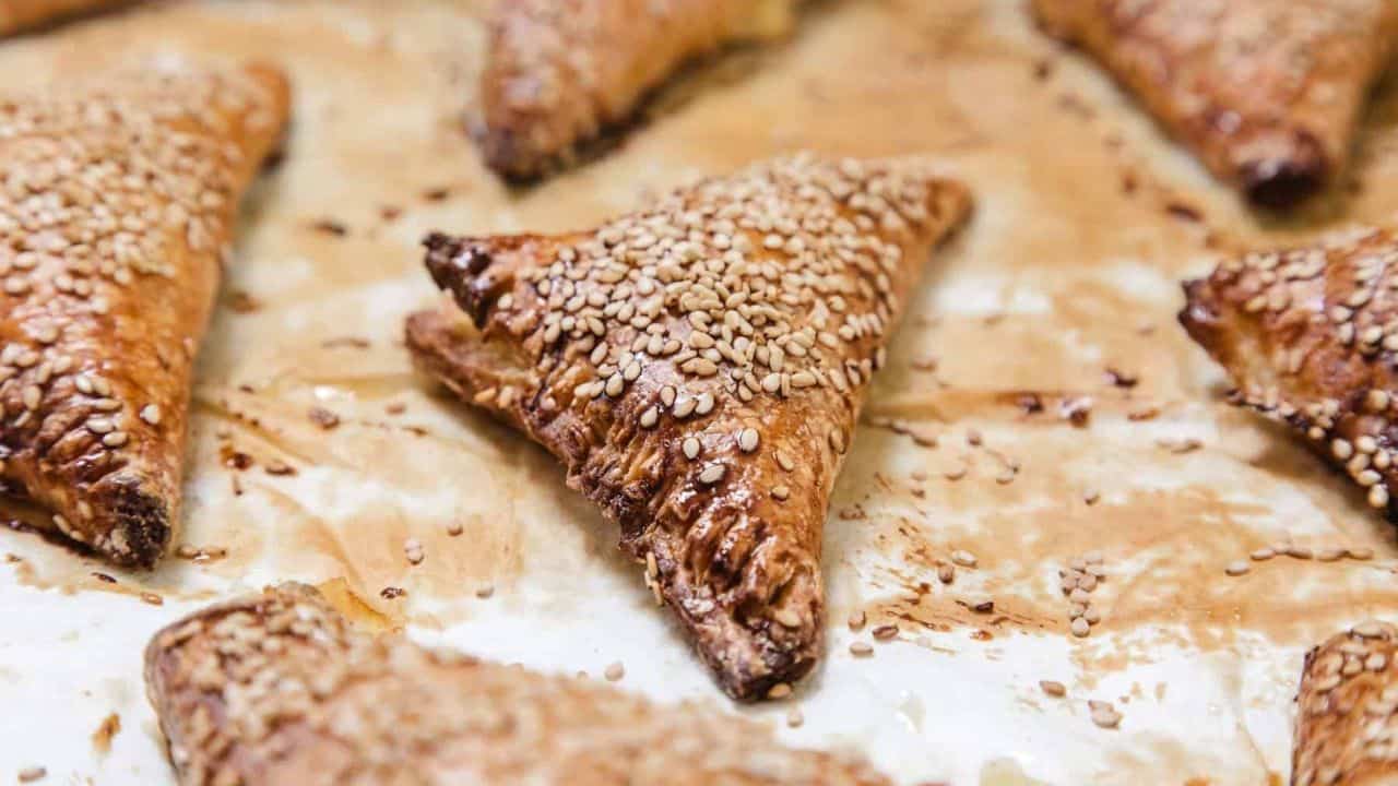 A close-up of golden, sesame-topped pastry triangles on a tray, served with fresh tomato halves and a sliced boiled egg.
