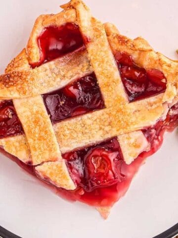 A slice of cherry pie with a lattice crust sits on a white plate, garnished with mint leaves, next to a fork and a red checkered napkin.