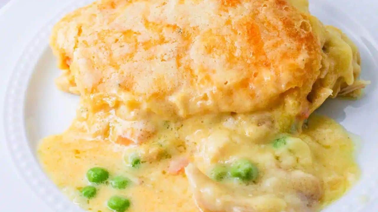 A serving of chicken pot pie with a golden-brown crust on a white plate. Visible peas and creamy filling are partially oozing out.
