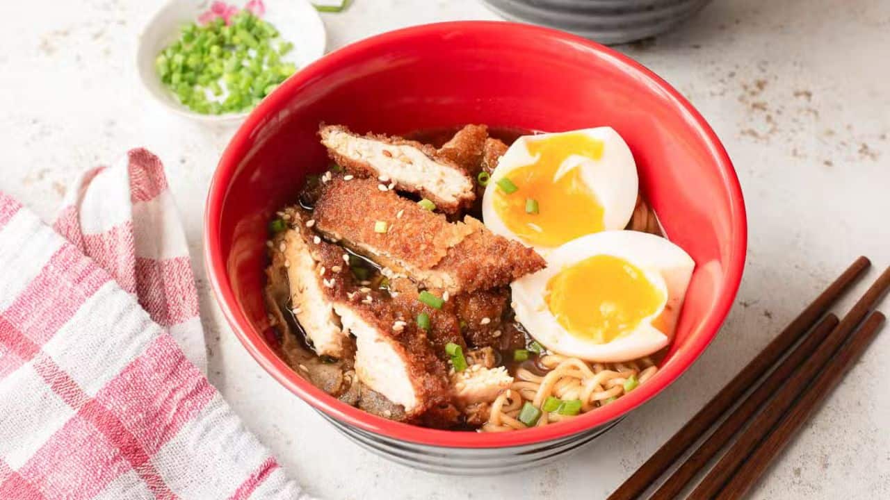 A red bowl filled with ramen noodles, topped with crispy breaded chicken slices, two halves of a soft-boiled egg, chopped green onions, and sesame seeds. Chopsticks and a small dish of green onions are beside the bowl.