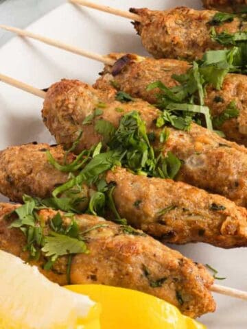 Five skewers of grilled kebabs garnished with chopped herbs are arranged on a white plate next to two lemon wedges.