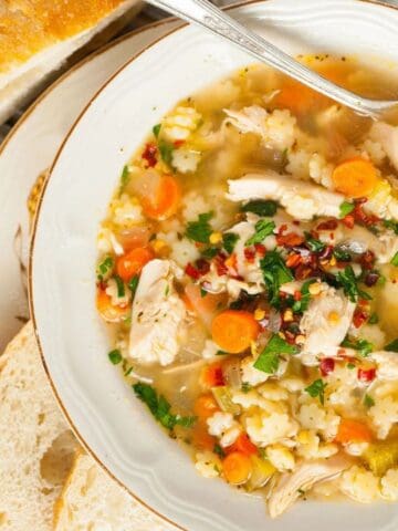 A bowl of chicken soup with carrots, pasta, and herbs, served with sliced bread on the side.