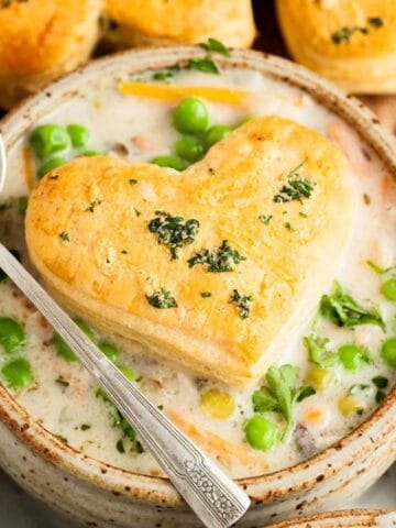 A bowl of creamy vegetable soup topped with a heart-shaped biscuit, garnished with parsley, and a spoon resting on the bowl.