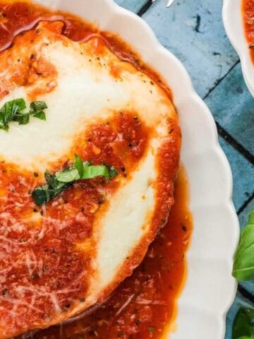 A close-up of eggplant parmesan topped with melted cheese, tomato sauce, grated cheese, and fresh basil, served in a white dish on a blue tiled surface.