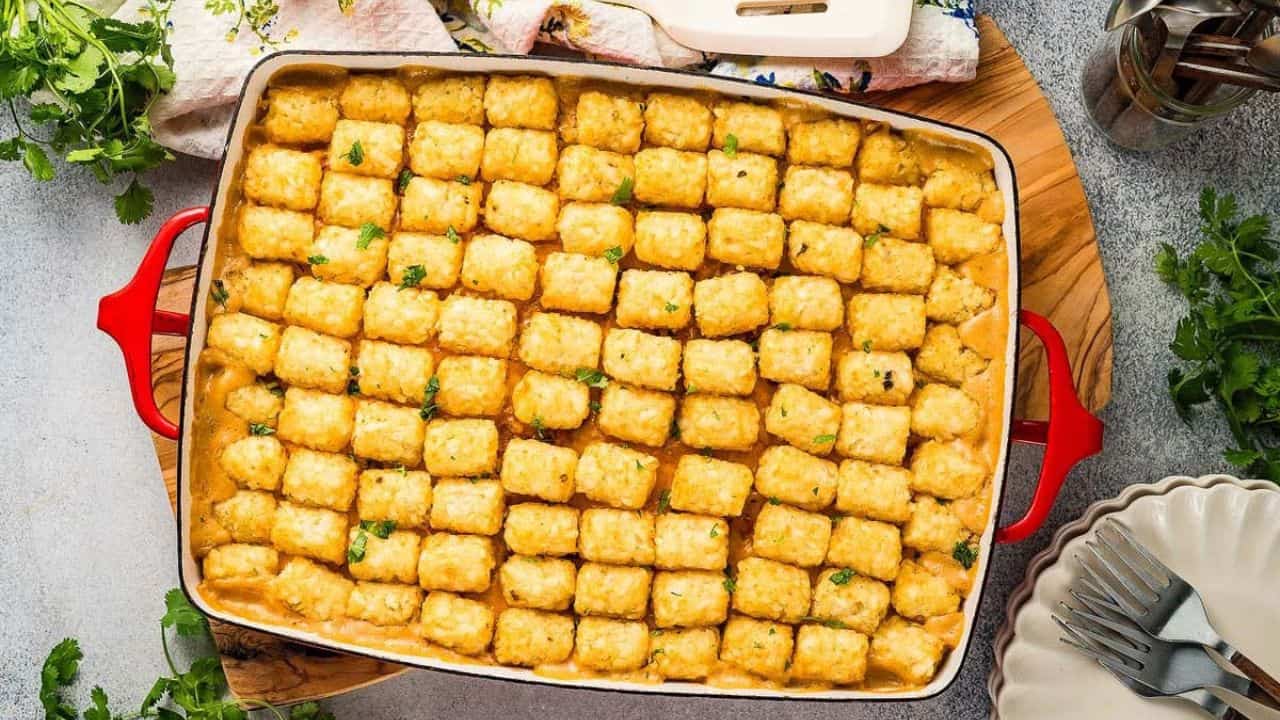 A casserole dish filled with neatly arranged tater tots on top of a baked, creamy filling, garnished with chopped herbs. Plates, utensils, and fresh herbs are nearby.