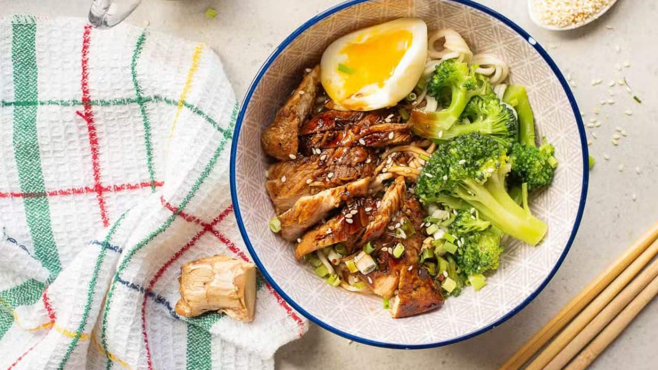 A hand uses chopsticks to pick up noodles from a bowl filled with ramen, broccoli, sliced egg, and garnished with sesame seeds and green onions. A towel and a piece of ginger are nearby on the countertop.